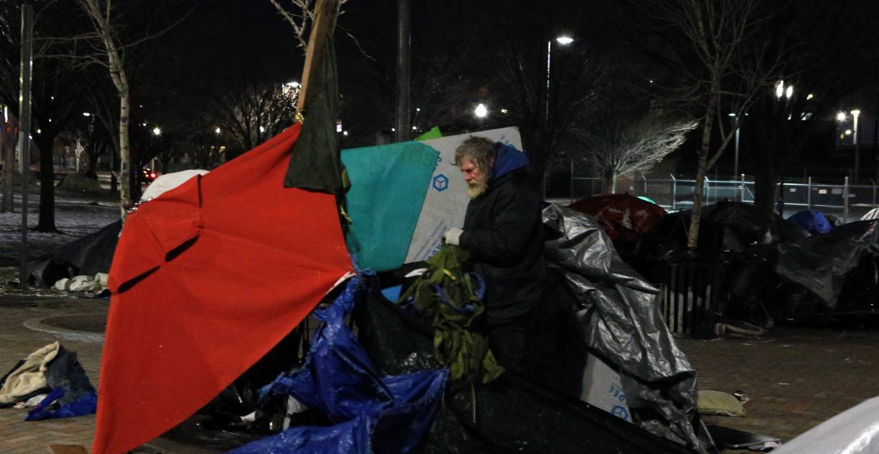 A person stands beside red and dark tarps and stacked belongings at night.