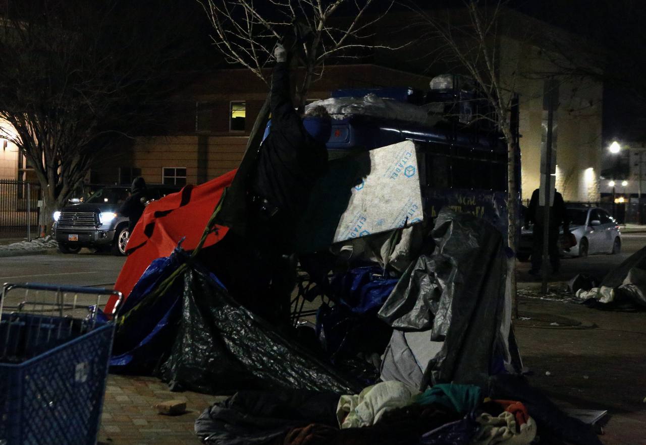Tarps, panels, bags, and a shopping cart beside a city sidewalk after dark.