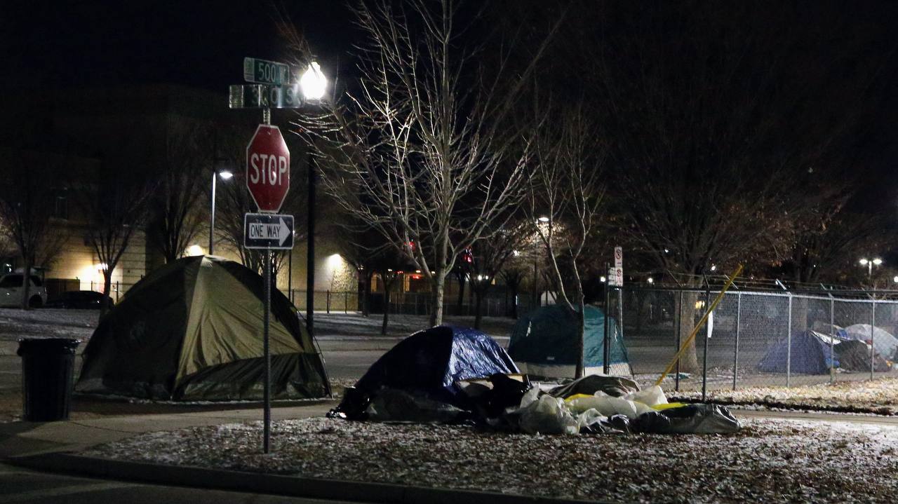 Tents stand along a sidewalk beneath streetlights, with patches of snow on the ground.