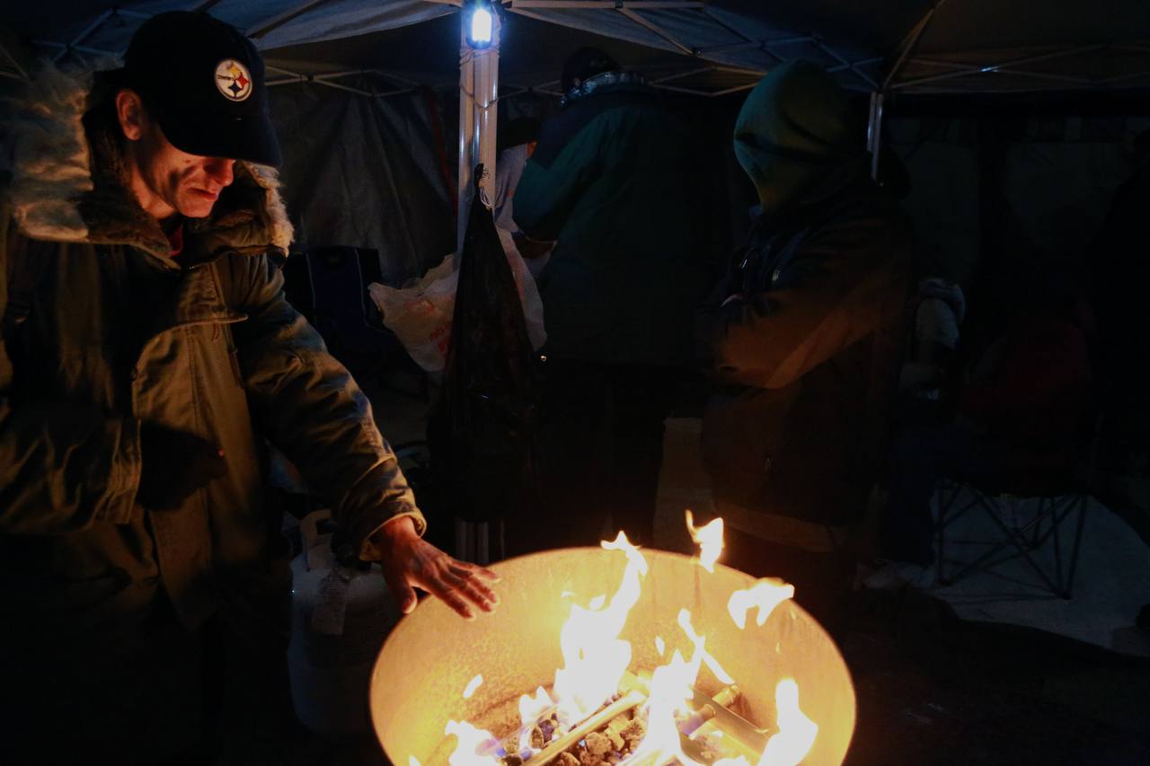 A person extends a hand toward a fire while others gather under a canopy.